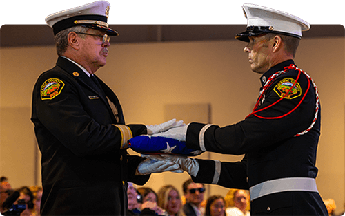 Two uniformed fire fighters participate in a flag presentation ceremony, with one handing a folded American flag to the other in front of an audience.